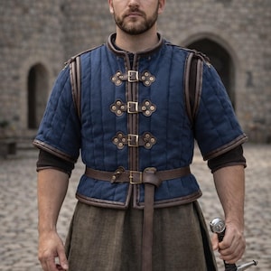 Puede incluir: Una persona con un atuendo de estilo medieval. El atuendo incluye un chaleco azul acolchado con detalles y hebillas de cuero marrón, un cinturón marrón y una falda marrón. La persona sostiene una espada.