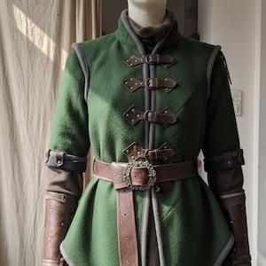 May include: A green wool jacket with brown leather accents and a matching belt. The jacket features decorative buckles and leather detailing on the sleeves. The garment is displayed on a mannequin, showcasing its medieval or fantasy-inspired design.