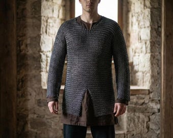 May include: A person wearing a chainmail shirt with long sleeves. The shirt is a dark gray color and has a split at the bottom. The person is wearing a brown shirt underneath. The background is a stone wall.