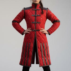 May include: A long, quilted red coat with brown leather accents and straps. The coat features a high collar, diamond stitching, and three buckle closures down the front. The shoulders and cuffs have dark gray leather details.