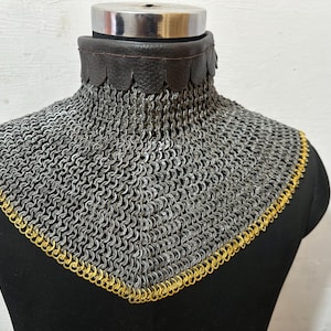 May include: A chainmail collar, likely for historical reenactment or costume use. The collar features a silver-colored chainmail body with a gold-colored trim along the bottom edge. A dark brown leather collar sits at the top.