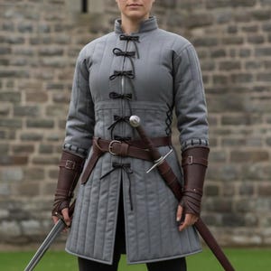 May include: A gray quilted tunic with black bow closures and a high collar. The tunic features a brown leather belt and matching arm bracers. A sword is sheathed at the waist. The garment is designed for historical reenactment or cosplay.
