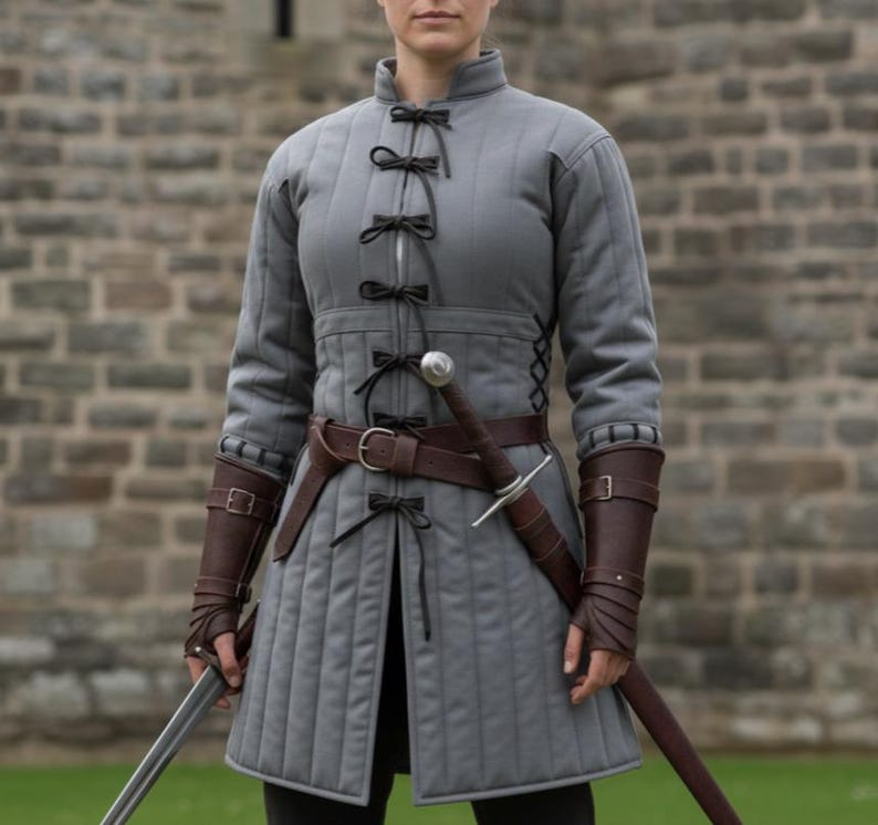 May include: A gray quilted tunic with black bow closures and a high collar. The tunic features a brown leather belt and matching arm bracers. A sword is sheathed at the waist. The garment is designed for historical reenactment or cosplay.