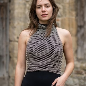 May include: A silver chainmail halter top with a high neckline. The top is made of small, interlocking metal rings. The model is wearing a black top and black pants. The background is a stone wall.