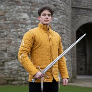 Könnte beinhalten: Senfgelber, gepolsterter Gambeson mit weißen Bindebändern, getragen über einem schwarzen Hemd. Der Gambeson hat einen hohen Kragen und wird mit einem langen, silbernen Schwert getragen. Der Hintergrund zeigt eine Steinmauer.