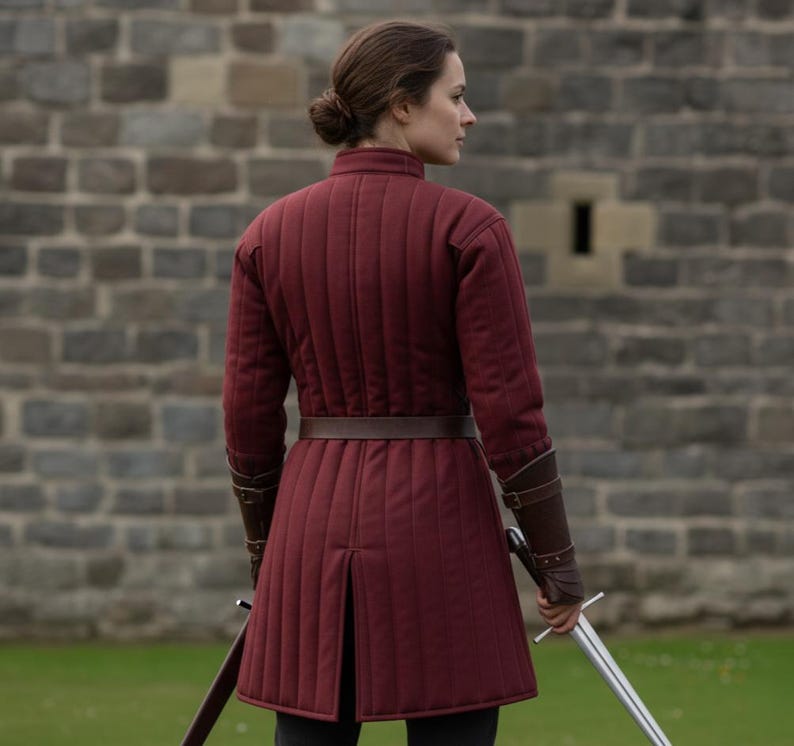 May include: A burgundy quilted tunic with a brown leather belt and matching wrist cuffs. The tunic has vertical stitching and a split at the hem. A sword is held at the side. The background is a stone wall.
