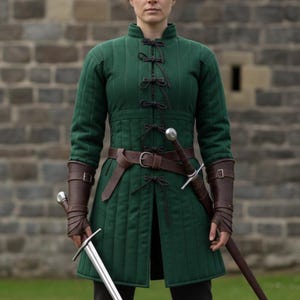 May include: A green quilted medieval tunic with black ties and a brown leather belt. The tunic is paired with brown leather arm guards and a sword. The background features a stone brick wall.