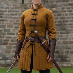 May include: Mustard-colored quilted medieval doublet with black ties and a brown leather belt. The outfit includes brown leather arm guards and a sword. The garment is worn over black pants.