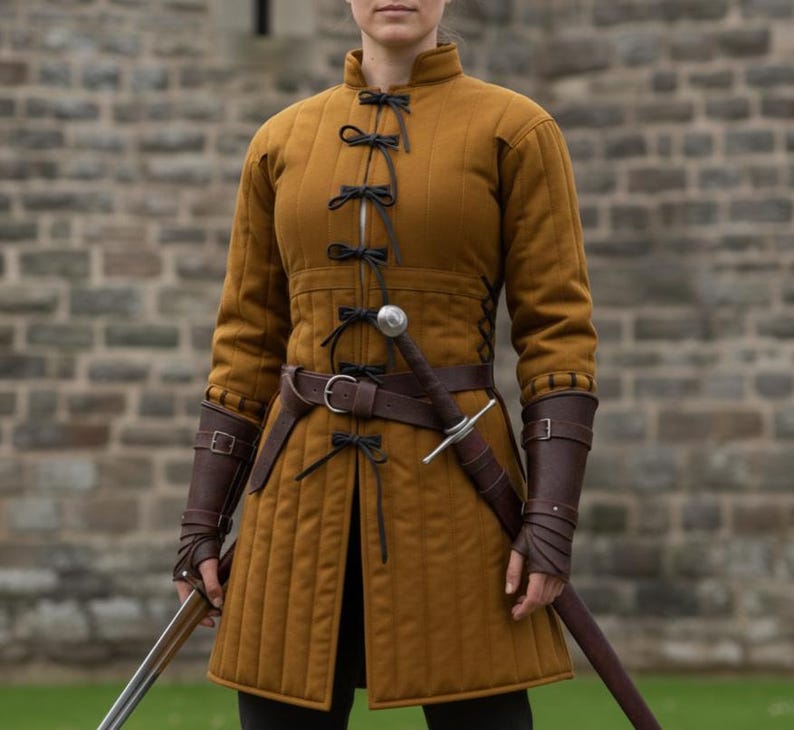 May include: Mustard-colored quilted medieval doublet with black ties and a brown leather belt. The outfit includes brown leather arm guards and a sword. The garment is worn over black pants.