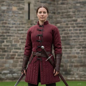 May include: A burgundy quilted tunic with black ties and a brown leather belt. The tunic has long sleeves and is paired with matching brown leather arm guards. A sword is sheathed at the waist.