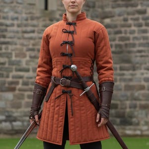 May include: An orange quilted medieval jacket with black ties and a brown leather belt. The jacket has long sleeves and is paired with brown leather arm guards. A sword is sheathed at the waist.