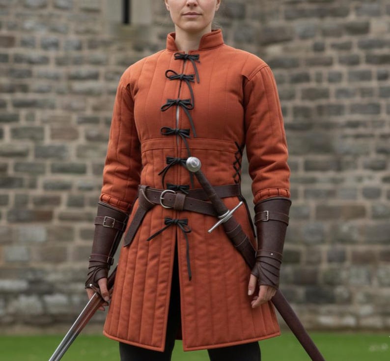 May include: An orange quilted medieval jacket with black ties and a brown leather belt. The jacket has long sleeves and is paired with brown leather arm guards. A sword is sheathed at the waist.