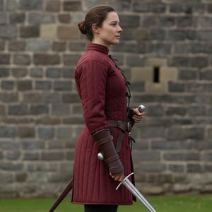 May include: A burgundy quilted gambeson with a high collar and long sleeves, worn with a brown leather belt and bracers. A silver sword is held at the ready. The background is a stone wall.