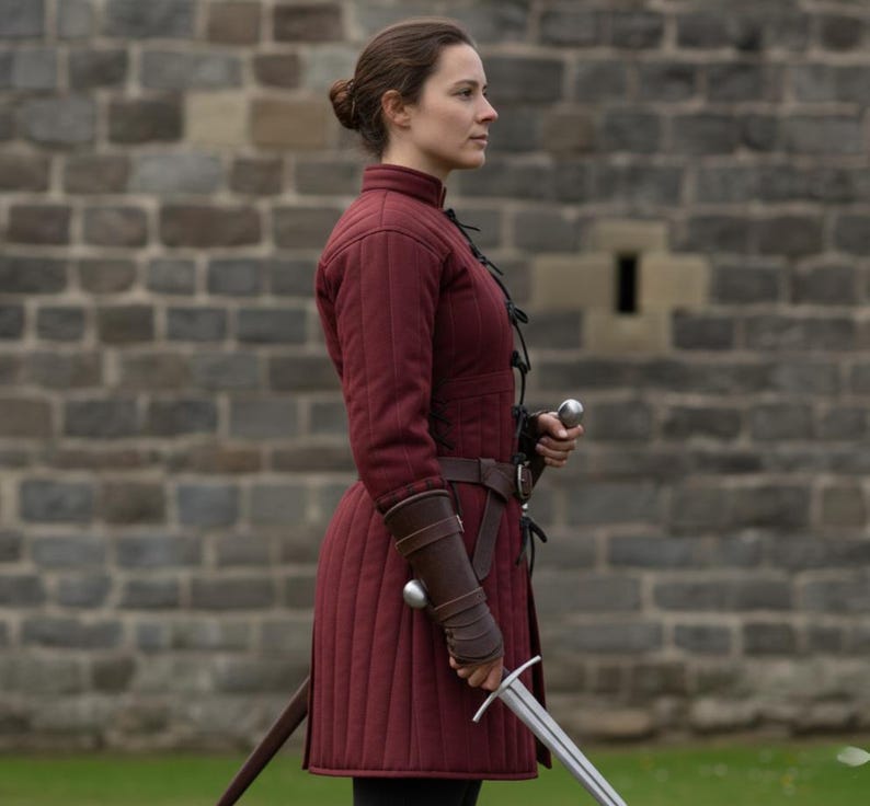 May include: A burgundy quilted gambeson with a high collar and long sleeves, worn with a brown leather belt and bracers. A silver sword is held at the ready. The background is a stone wall.