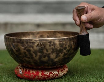 May include: A bronze-colored metal singing bowl with a mottled, dark patina and a circular symbol etched on the side. The bowl rests on a round, red fabric cushion featuring gold floral patterns, accompanied by a wooden mallet with a black tip.