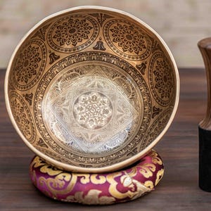 May include: A golden singing bowl with detailed etched patterns, on a patterned cushion. A wooden mallet with a black base is next to the bowl. The bowl's design includes floral and geometric motifs.