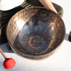 Puede incluir: Un cuenco tibetano de bronce texturizado con un mazo de madera y un percutor rojo. El cuenco es redondo y tiene un interior oscuro y reflectante. También hay un mazo de madera más pequeño con base negra.