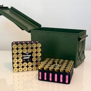 Puede incluir: Una caja de munición de metal verde oliva con la tapa abierta, acompañada de dos soportes de plástico negro que contienen cartuchos de escopeta calibre 12 de color rosa con bases de latón. El texto 12 Guage está impreso en blanco.