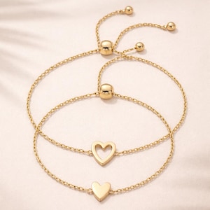 Puede incluir: Dos pulseras doradas con cierres de cadena ajustables. Una pulsera presenta un corazón calado y la otra un dije de corazón macizo. Las pulseras se exhiben sobre una superficie suave y clara.