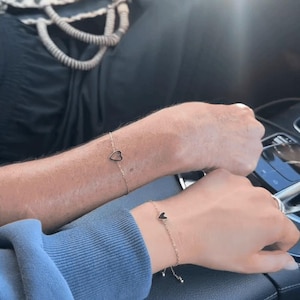 May include: Two gold-colored chain bracelets, each featuring a small black heart charm. The bracelets are worn on wrists, with one arm in a blue sweater and the other in a black top. The bracelets are delicate and simple.