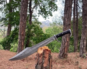 May include: A large, ornate knife with a patterned blade and a black handle rests on a tree stump. The blade has a unique, swirling design. The handle features a curved guard. The background includes trees and foliage, suggesting an outdoor setting.