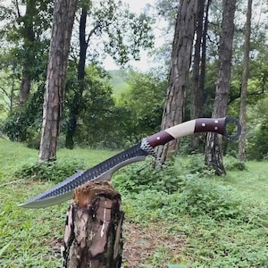 May include: A large, curved blade knife with a dark, serrated edge and a handle featuring a white, brown, and red design. The knife is resting on a tree stump in a forest setting, showcasing its craftsmanship and design.