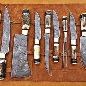 Op de afbeelding: Een set Damascus stalen messen met bot- en geweihandvatten, gepresenteerd in een bruine leren rol. De messen hebben een uniek wervelpatroon. De set bevat diverse messen en een trancheervork.