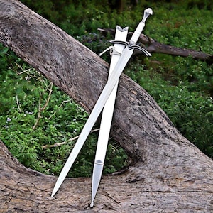 May include: A silver sword with a white handle and a matching sheath rests on a weathered log. The sword features a cross-shaped hilt and a long, pointed blade. The background is a lush green with foliage.