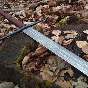 May include: A long, silver sword with a patterned blade and a brown leather-wrapped handle. The sword rests on a weathered tree stump surrounded by fallen leaves, evoking a medieval or fantasy theme.