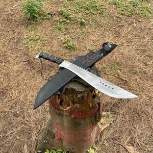 May include: A large, silver machete with a black handle and sheath. The blade has a curved edge and several holes. The machete rests on a tree stump. The blade is approximately 45 cm long.