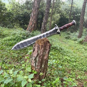 Puede incluir: Una espada grande, forjada a mano, con una hoja oscura y texturizada y un mango de madera. La espada descansa sobre un tocón de árbol en un entorno forestal, mostrando su artesanía y diseño. La hoja mide aproximadamente 76 cm de largo.