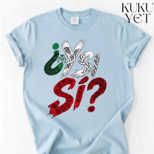 Op de afbeelding: Een lichtblauw t-shirt met een grafisch ontwerp met de tekst ¿Y SI? in groen, wit en rood. De letters bevatten ingewikkelde patronen, waaronder een adelaar, veren en geometrische vormen. De merknaam KUKU YET staat in het zwart rechtsboven afgedrukt.