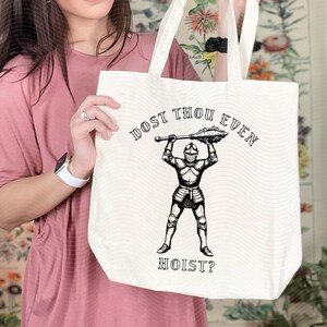 May include: Cream-colored tote bag with black text and graphic. The text reads "DOST THOU EVEN HOIST?" above and below a black illustration of a knight holding a mace. The bag has white handles.