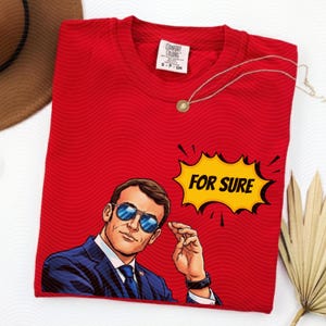 以下が含まれることがあります： サングラスをかけた男性のポップアート風イラストと、黄色の吹き出しに「FOR SURE」の文字が入った赤いTシャツ。 金色のネックレスがシャツにかけられています。 シャツにはComfort Colorsのタグが付いています。