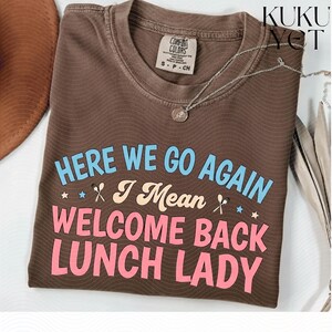 Peut inclure: Un t-shirt marron à manches courtes avec un col rond. Le t-shirt présente le texte "HERE WE GO AGAIN I Mean WELCOME BACK LUNCH LADY" en bleu clair, crème et rose, agrémenté de petites étoiles et d'icônes de cuillères. Un collier argenté est posé dessus.