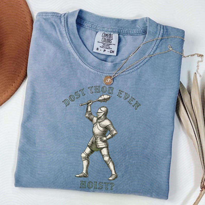 May include: A light blue Comfort Colors t-shirt with the text "DOST THOU EVEN HOIST?" and an illustration of a knight. A gold necklace is draped across the shirt. The shirt is folded on a white surface.