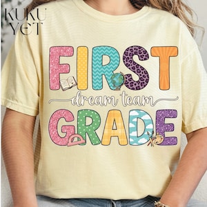 Peut inclure: Un t-shirt à manches courtes jaune pâle avec le texte KUKU YET dans le coin supérieur gauche. Le motif central affiche les mots FIRST GRADE en grandes lettres colorées et à motifs, avec le texte dream team écrit en cursif au milieu.