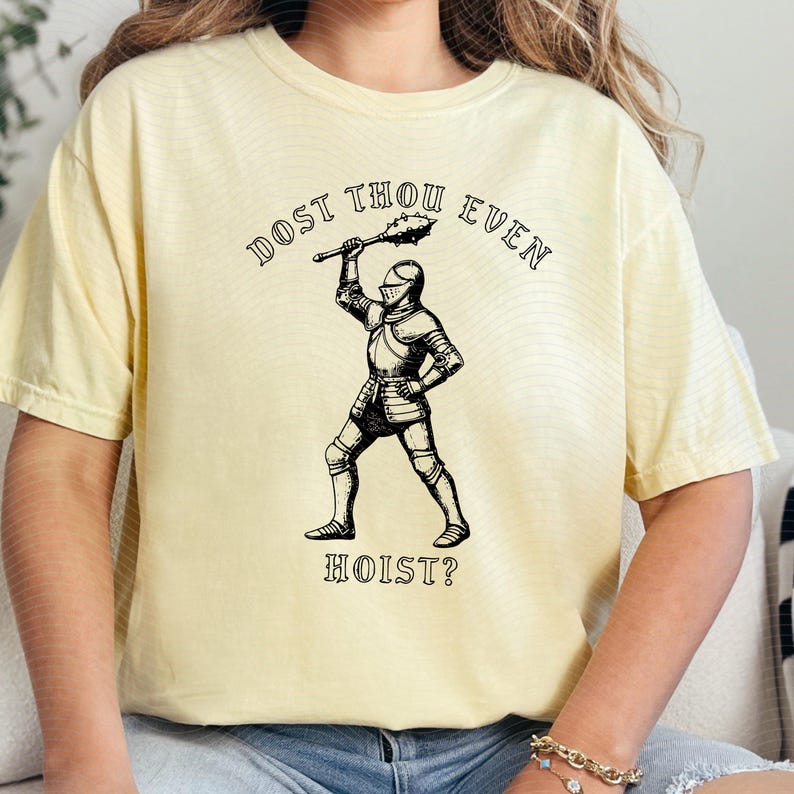 May include: A light yellow t-shirt featuring a black and white illustration of a knight holding a mace. The text "DOST THOU EVEN HOIST?" is arched above and below the knight. The shirt has short sleeves and a relaxed fit.