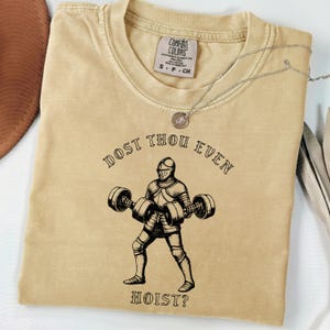 May include: A tan Comfort Colors t-shirt with the text "DOST THOU EVEN HOIST?" above and below a black illustration of a knight lifting weights. A silver necklace with a pendant is draped across the shirt.