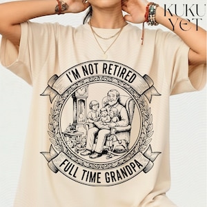 Peut inclure: T-shirt beige avec une illustration en noir et blanc d'un grand-père lisant à trois enfants. Le texte dit "I'M NOT RETIRED FULL TIME GRANDPA" dans un design circulaire avec une bannière.