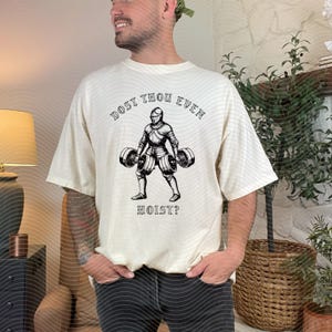 May include: Cream-colored t-shirt with a black graphic of a knight lifting barbells. The text "DOST THOU EVEN" arches above the knight, and "HOIST?" is below. The shirt is a relaxed fit.