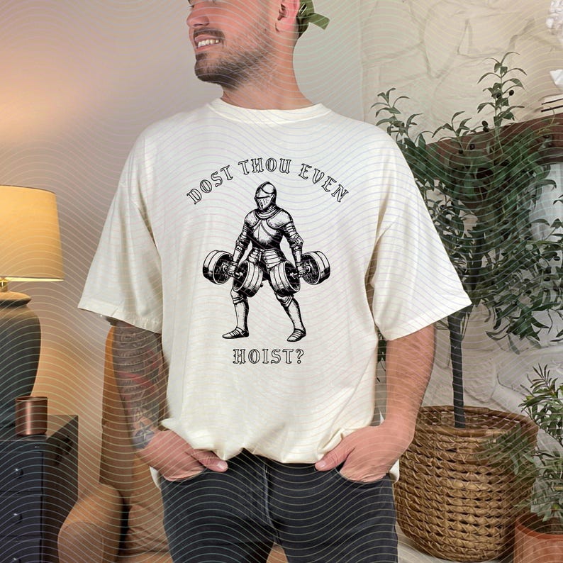 May include: Cream-colored t-shirt with a black graphic of a knight lifting barbells. The text "DOST THOU EVEN" arches above the knight, and "HOIST?" is below. The shirt is a relaxed fit.