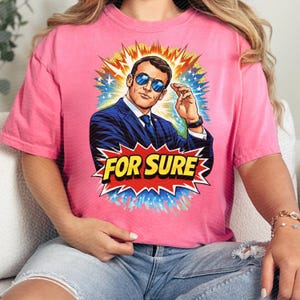 以下が含まれることがあります： ピンク色のTシャツ。スーツとサングラスをかけた男性のポップアート風イラストが特徴です。画像は黄色、オレンジ、青、白のバーストで囲まれています。「FOR SURE」の文字が赤と黄色のスターバーストで表示されています。