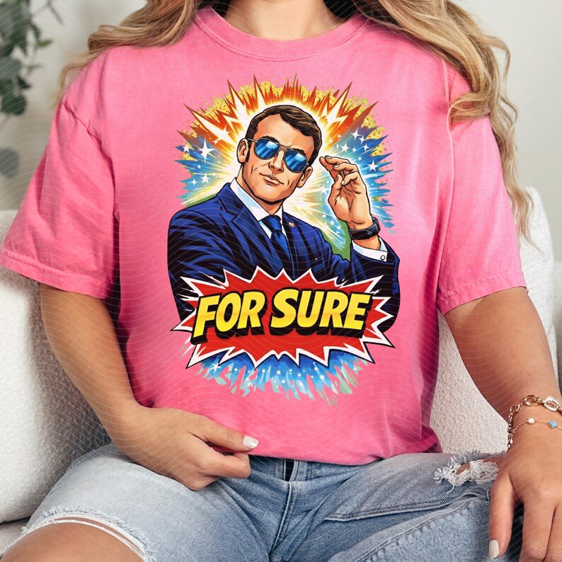 以下が含まれることがあります： ピンク色のTシャツ。スーツとサングラスをかけた男性のポップアート風イラストが特徴です。画像は黄色、オレンジ、青、白のバーストで囲まれています。「FOR SURE」の文字が赤と黄色のスターバーストで表示されています。