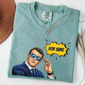 以下が含まれることがあります： 水色のComfort Colors Tシャツ。サングラスとスーツを着た男性の漫画イラストが描かれています。「FOR SURE」と書かれた黄色の吹き出しがあります。シルバーのネックレスがTシャツの上にかけられています。