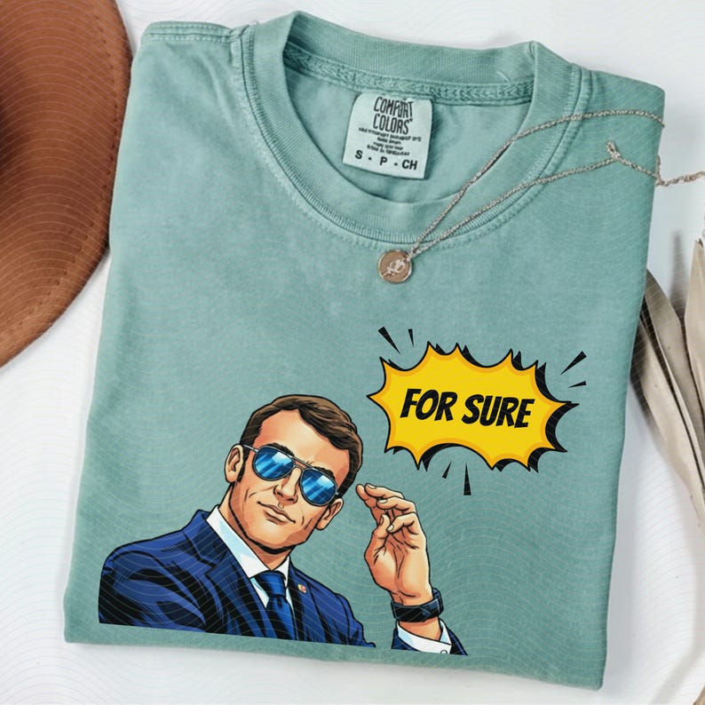 以下が含まれることがあります： 水色のComfort Colors Tシャツ。サングラスとスーツを着た男性の漫画イラストが描かれています。「FOR SURE」と書かれた黄色の吹き出しがあります。シルバーのネックレスがTシャツの上にかけられています。