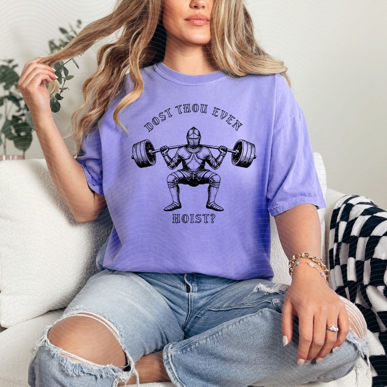 May include: A light purple t-shirt with a black graphic of a knight lifting a barbell. The text "DOST THOU EVEN HOIST?" is above and below the knight. The shirt is made of a soft material.