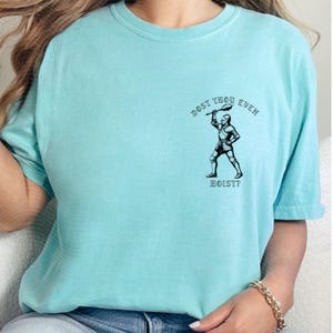 May include: A light blue t-shirt featuring a black and white illustration of a knight holding a weapon. The text "DOST THOU EVEN HOIST?" is printed above and below the knight image. The shirt has short sleeves.