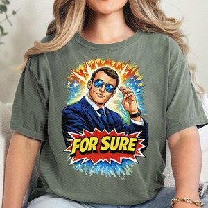 以下が含まれることがあります： オリーブグリーンのTシャツ。スーツとサングラスをかけた男性のコミックスタイルのイラストが描かれています。画像は、黄色、青、白の星のバーストに囲まれ、赤と黄色のバナーに「FOR SURE」という言葉が書かれています。