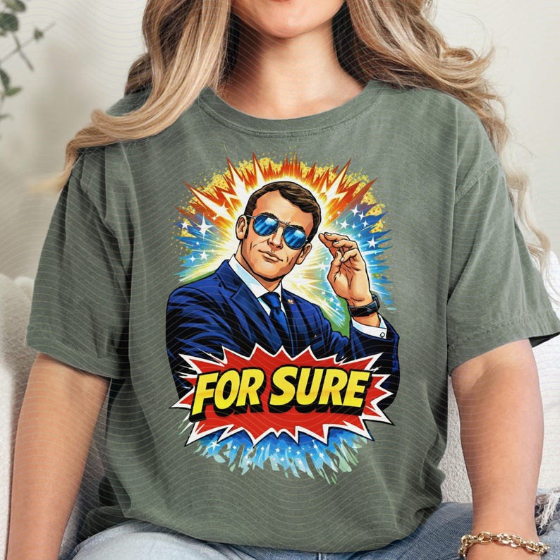 以下が含まれることがあります： オリーブグリーンのTシャツ。スーツとサングラスをかけた男性のコミックスタイルのイラストが描かれています。画像は、黄色、青、白の星のバーストに囲まれ、赤と黄色のバナーに「FOR SURE」という言葉が書かれています。